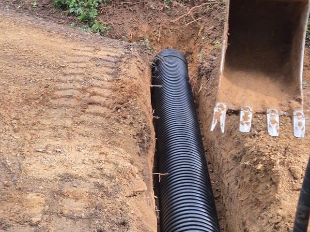 Black corrugated drainage pipe laid in a freshly dug trench, with an excavator bucket alongside