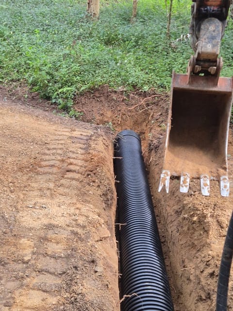 Black corrugated drainage pipe laid in a freshly dug trench, with an excavator bucket alongside