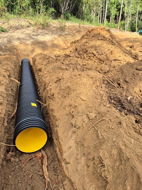 Large black corrugated drainage pipe laid in a freshly dug trench, with the spoil heaped alongside