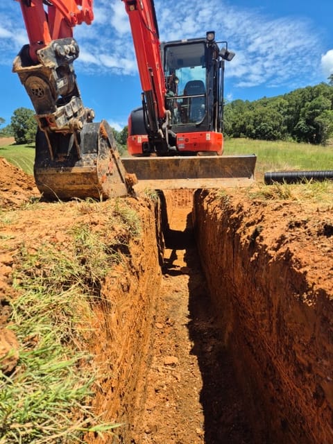 Orange Kubota excavator at the end of a deep, clean-walled trench in red soil, with drainage pipe ready beside it