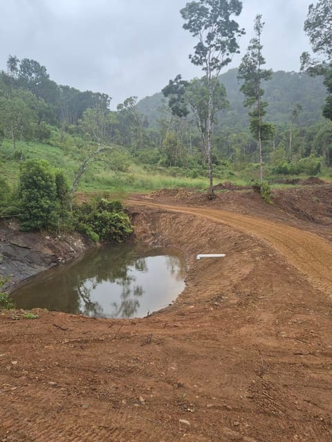 Newly dug farm dam holding water beside a freshly formed dirt track, with forested hills behind